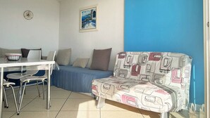 A room with a blue wall, a sofa with a patterned cover, a dining table with chairs, a clock, and a framed picture on the wall. - Seafront studio with private parking - Le Lavandou (Le Lavandou)