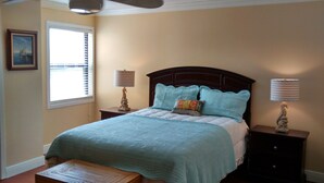 A bedroom with a bed, two bedside tables, a window with blinds, a painting on the wall, and a ceiling fan. - Beauvoir (Beautiful View) (Biloxi)