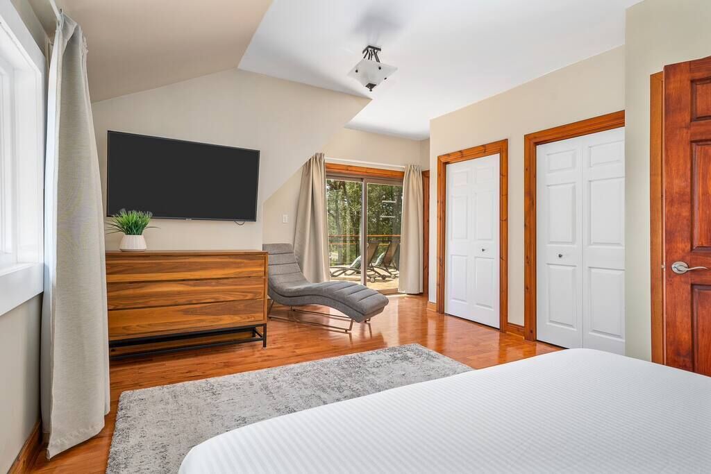 A bedroom with a bed, a wooden dresser, a chair, a TV, and a sliding glass door leading to a balcony.