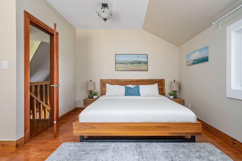Une chambre avec un grand lit, une tête de lit en bois, deux tables de chevet avec des lampes, un tableau au mur et une fenêtre donnant sur un beau paysage.