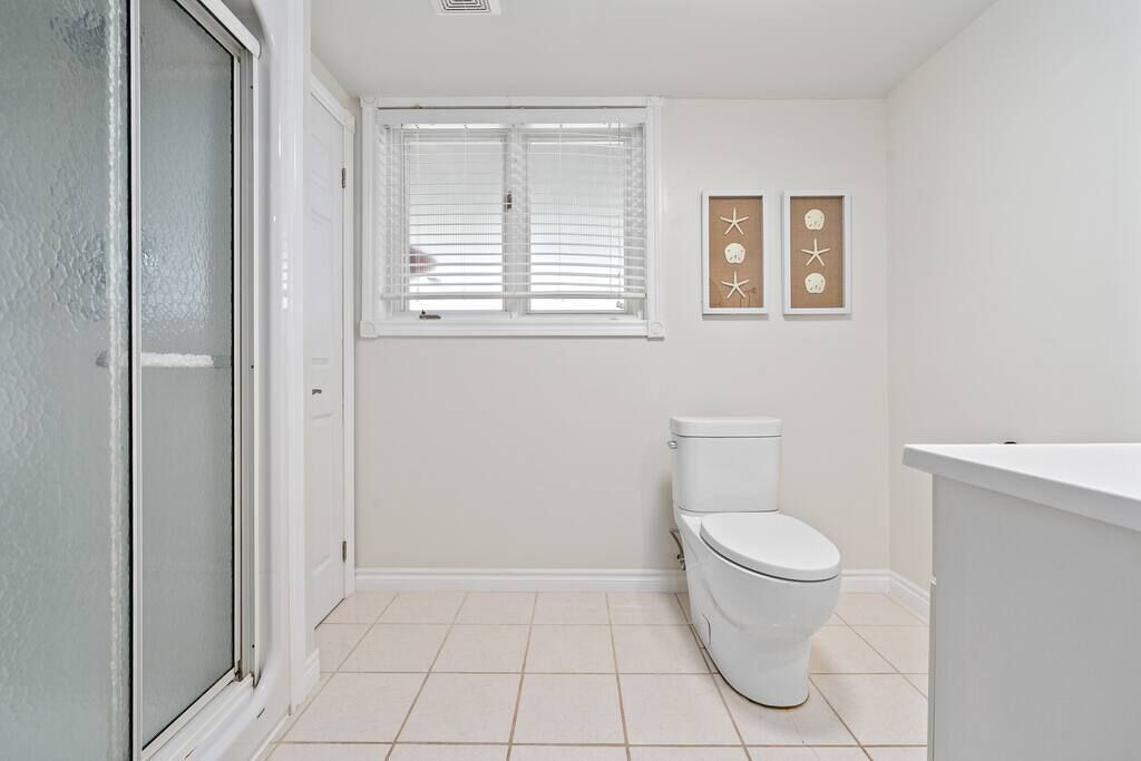 Une salle de bain avec des toilettes, une fenêtre avec des stores et deux tableaux encadrés au mur.