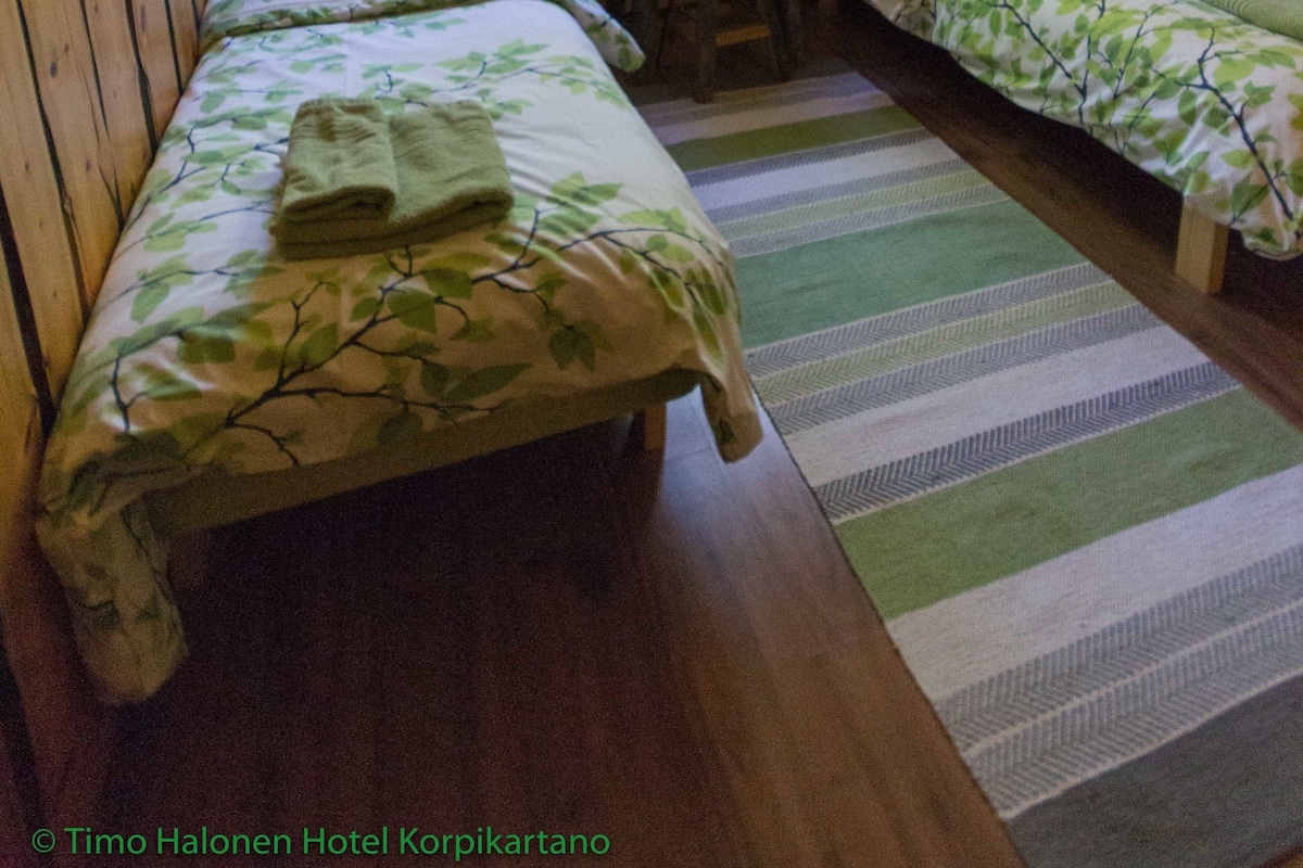 A room with two single beds, a wooden ceiling, a window with curtains, a desk with a chair, and a striped rug.