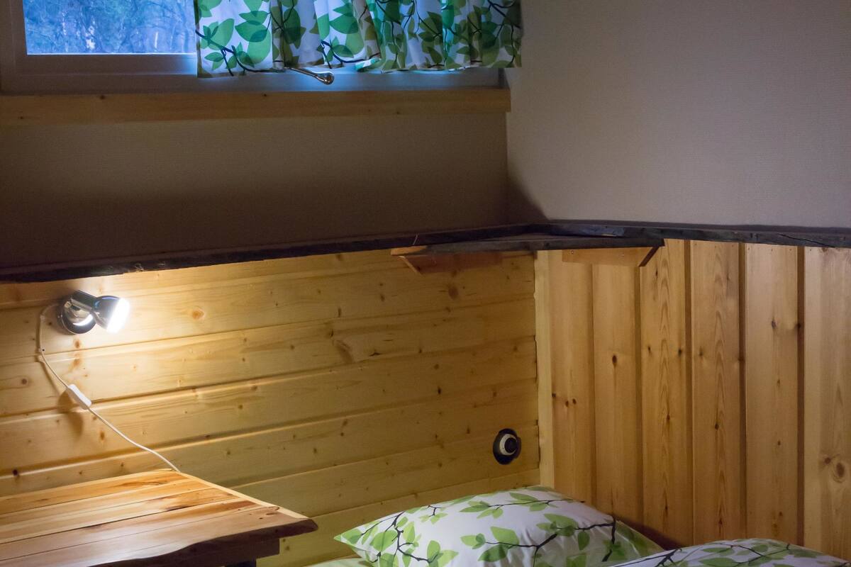 A small, cozy room with a wooden bunk bed, a desk with a chair, and a window with green leaf-patterned curtains.