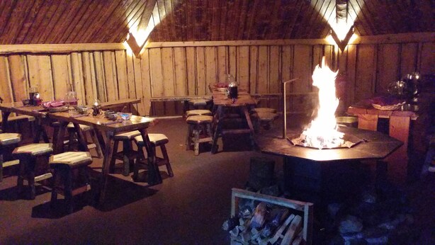Ein rustikaler Essbereich mit Holztischen und -stühlen, einer zentralen Feuerstelle und einer Holzkονstruktion an der Decke.