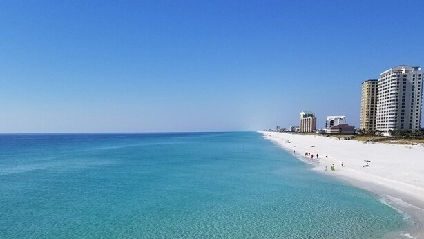 Ein Strand mit klarem türkisblauem Wasser, weißem Sand und einigen Gebäuden im Hintergrund.