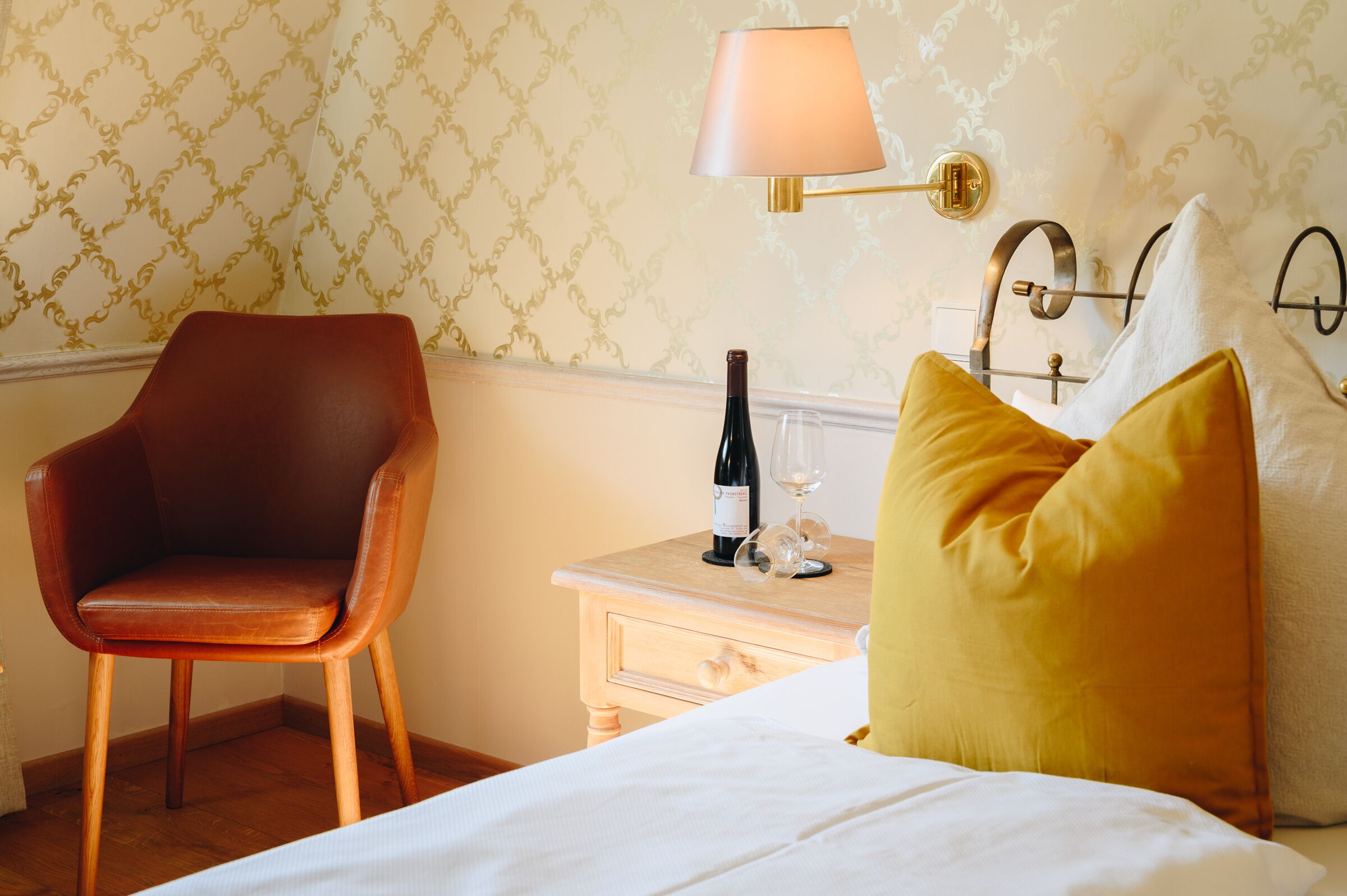 Ein Hotelzimmer mit einem Holzstuhl, einem Nachttisch mit einer Weinflasche und einem Glas sowie einer Lampe.