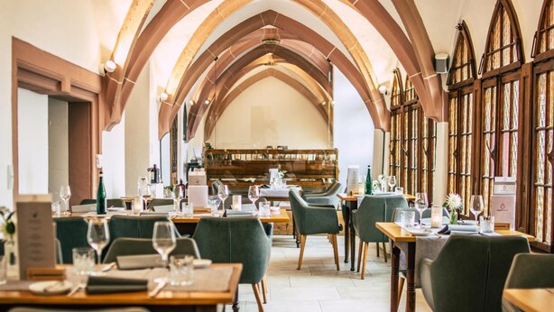 Un comedor con mesas dispuestas para dos, sillas de madera y un techo alto con arcos y ventanas de vitrales.