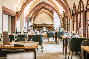 Een eetgedeelte met tafels voor twee personen, houten stoelen en een hoge gewelfde plafond met glas-in-loodramen.