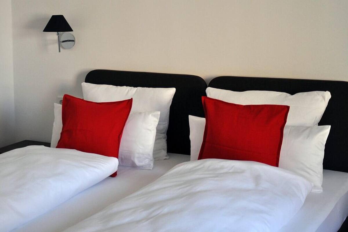Une chambre d’hôtel avec un lit doté d’une literie blanche et rouge, d’une tête de lit noire et d’une lampe fixée au mur.