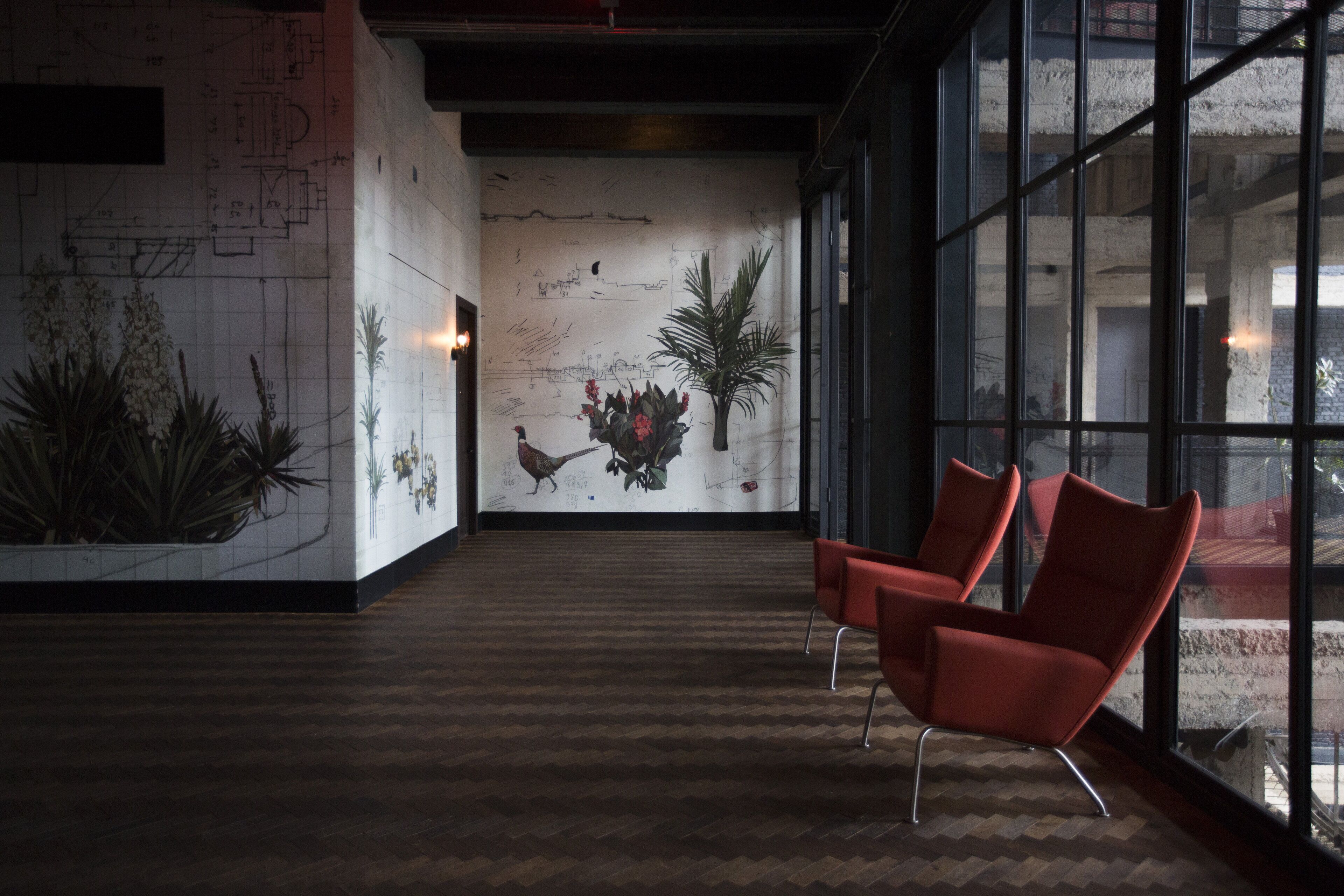 A hallway with a wooden floor, red chairs, a large window, and a wall with architectural drawings and plants.