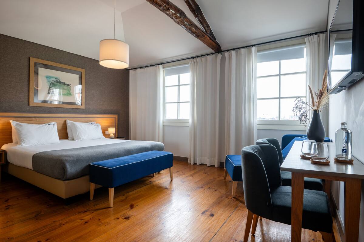 A modern hotel room with a large bed, a desk with chairs, a vase with dried plants, and a window with curtains.