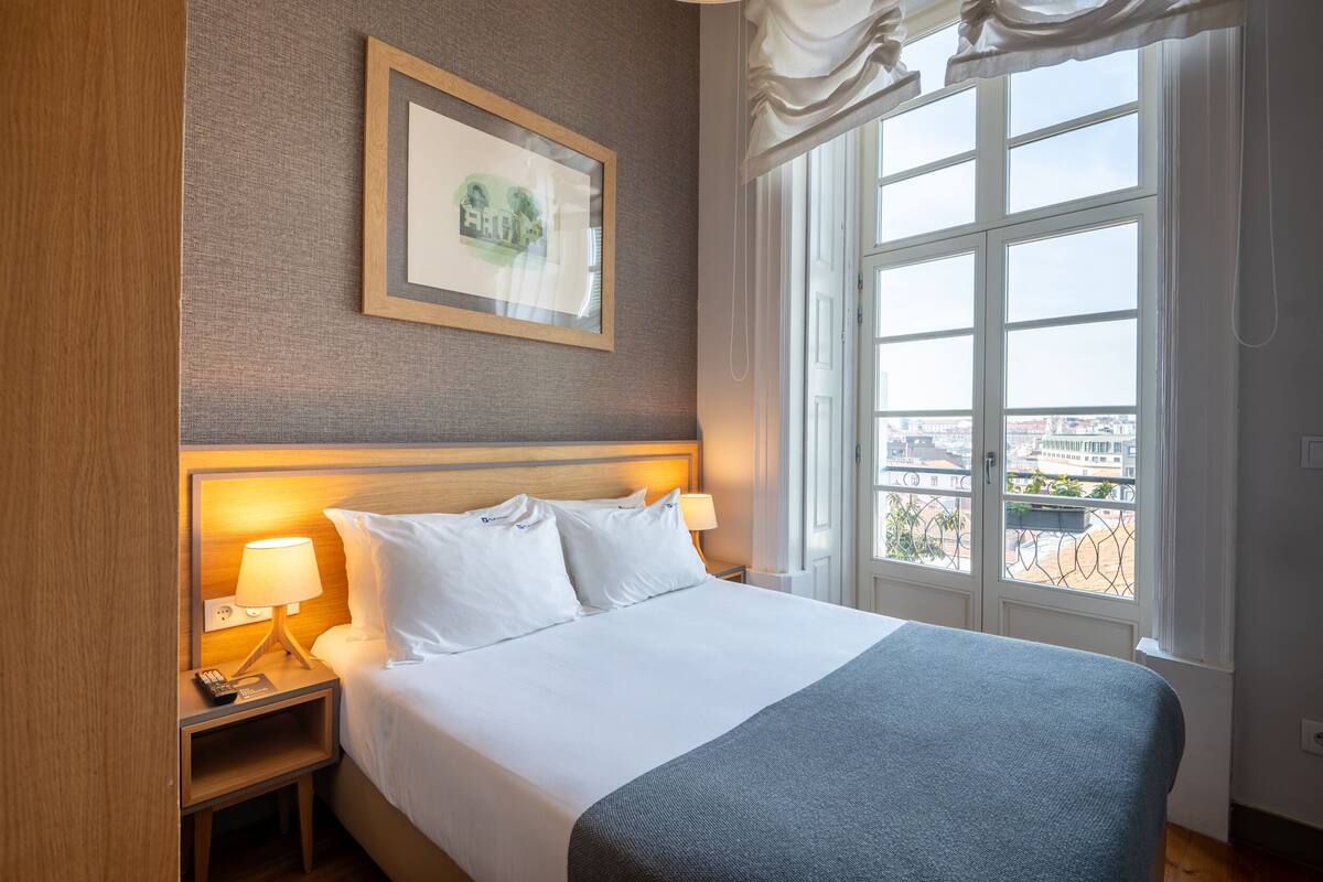 A hotel room with a large bed, two bedside tables with lamps, a framed picture on the wall, and a large window with a city view.