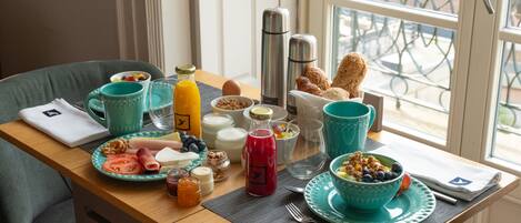 A breakfast table with various food items, a coffee maker, and a window with a view of the street.