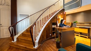 A wooden staircase with white railings leading to a second floor. A person is seated at a desk with a laptop, and there is a plant on the floor. - Santa Catarina FLH Suites (Porto)