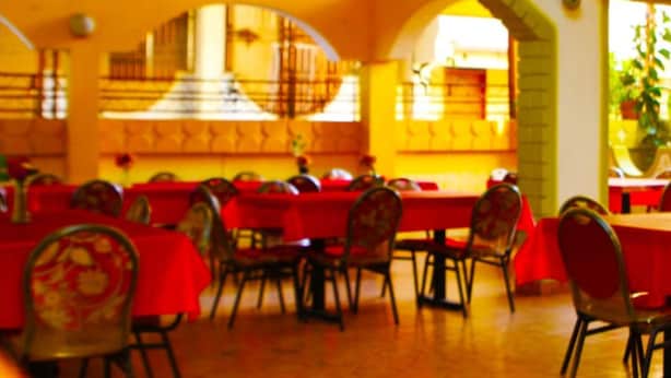 A restaurant with red tables and chairs, yellow walls, and a high ceiling with a hanging light fixture. - Hotel Le Meridien (Les Cayes)