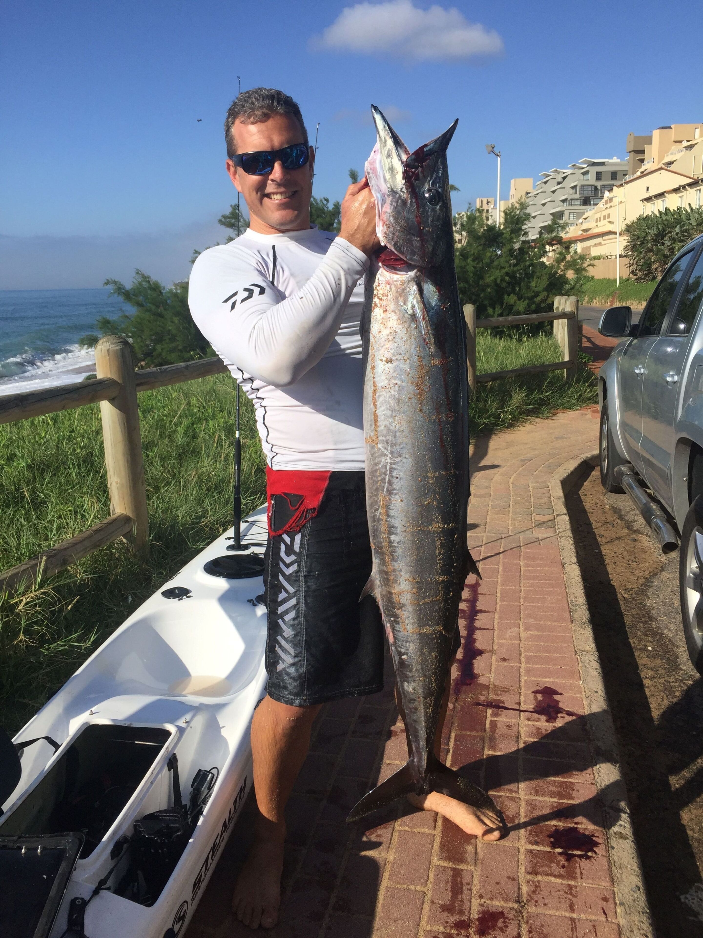 A person holding a large fish next to a kayak and a car.