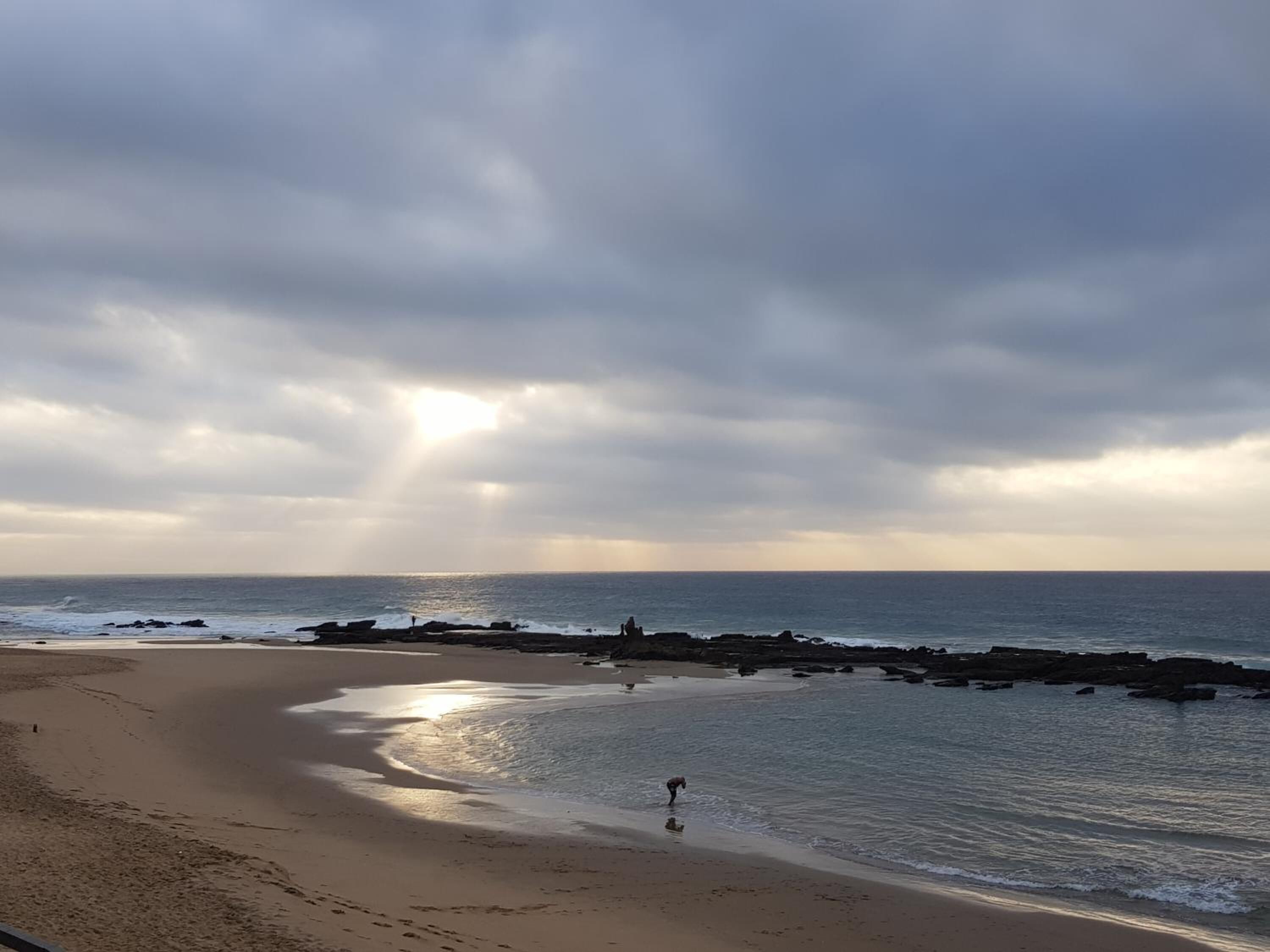 A beach with a curved shoreline, rocky outcrops, and a person walking along the water's edge.