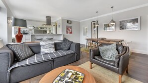 Ein modernes Wohnzimmer mit einem grauen Sofa, einem braunen Ledersessel und einem Couchtisch aus Holz. Es gibt einen Essbereich mit einem Holztisch und Stühlen. Der Raum hat weiße Wände und ein großes Fenster.