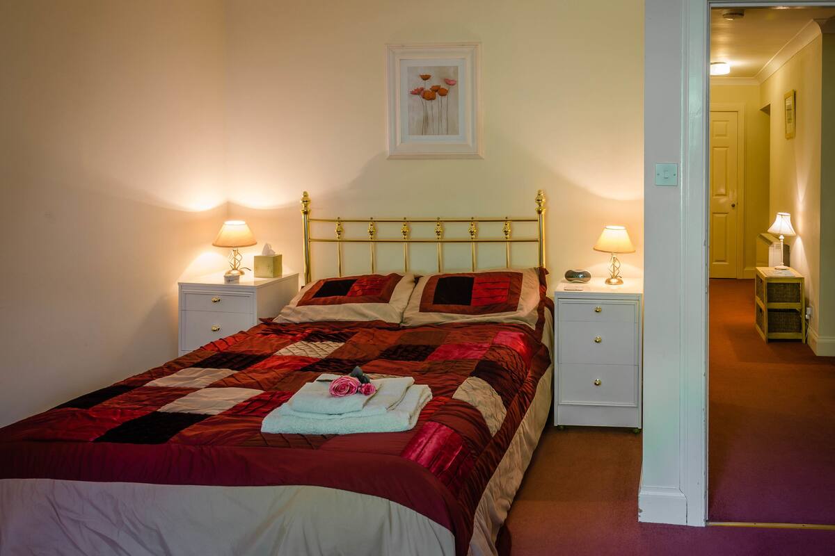 A neatly made bed with a red and black quilt, two bedside tables with lamps, a framed picture on the wall, and a door leading to another room.