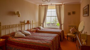 A hotel room with two beds, a sitting area with a chair and a table, a window with curtains, and a wall-mounted lamp.