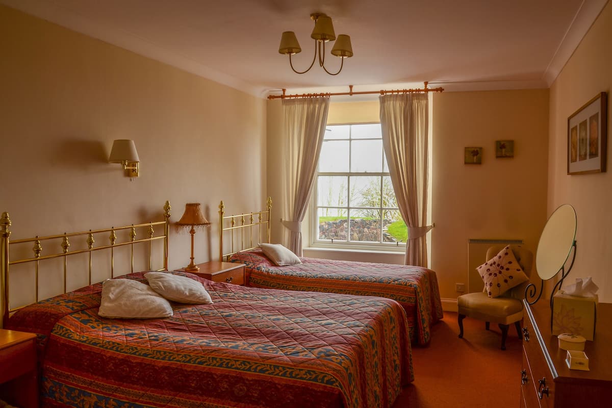 A hotel room with two beds, a sitting area with a chair and a table, a window with curtains, and a wall-mounted lamp.