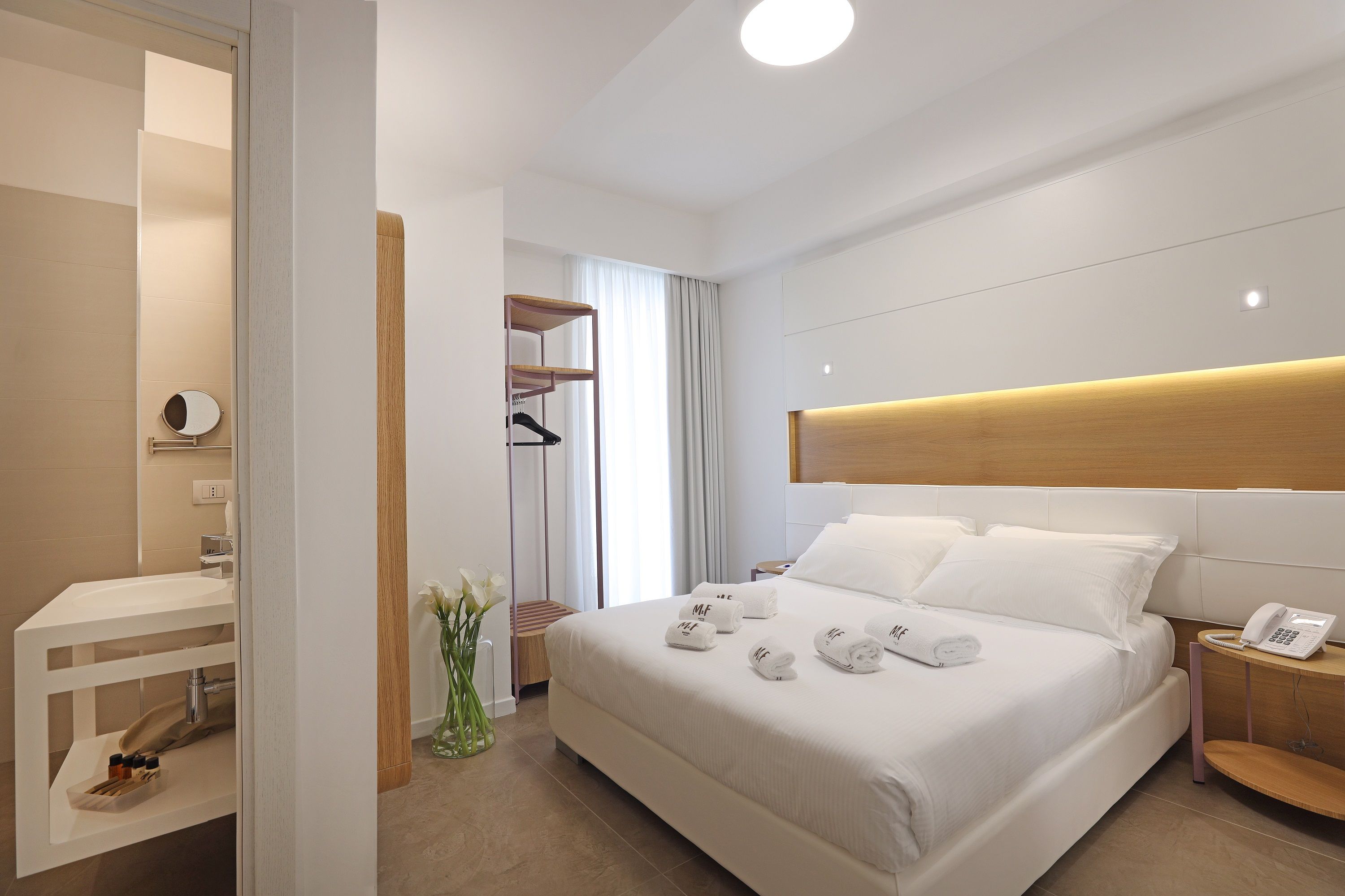 A modern hotel room with a large bed, a bedside table with a phone, and a small desk with a mirror and toiletries.