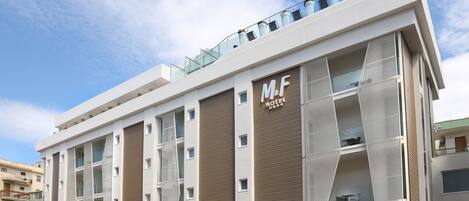 Un moderno edificio del hotel M&F con fachada plana, ventanas grandes y un jardín en la azotea.