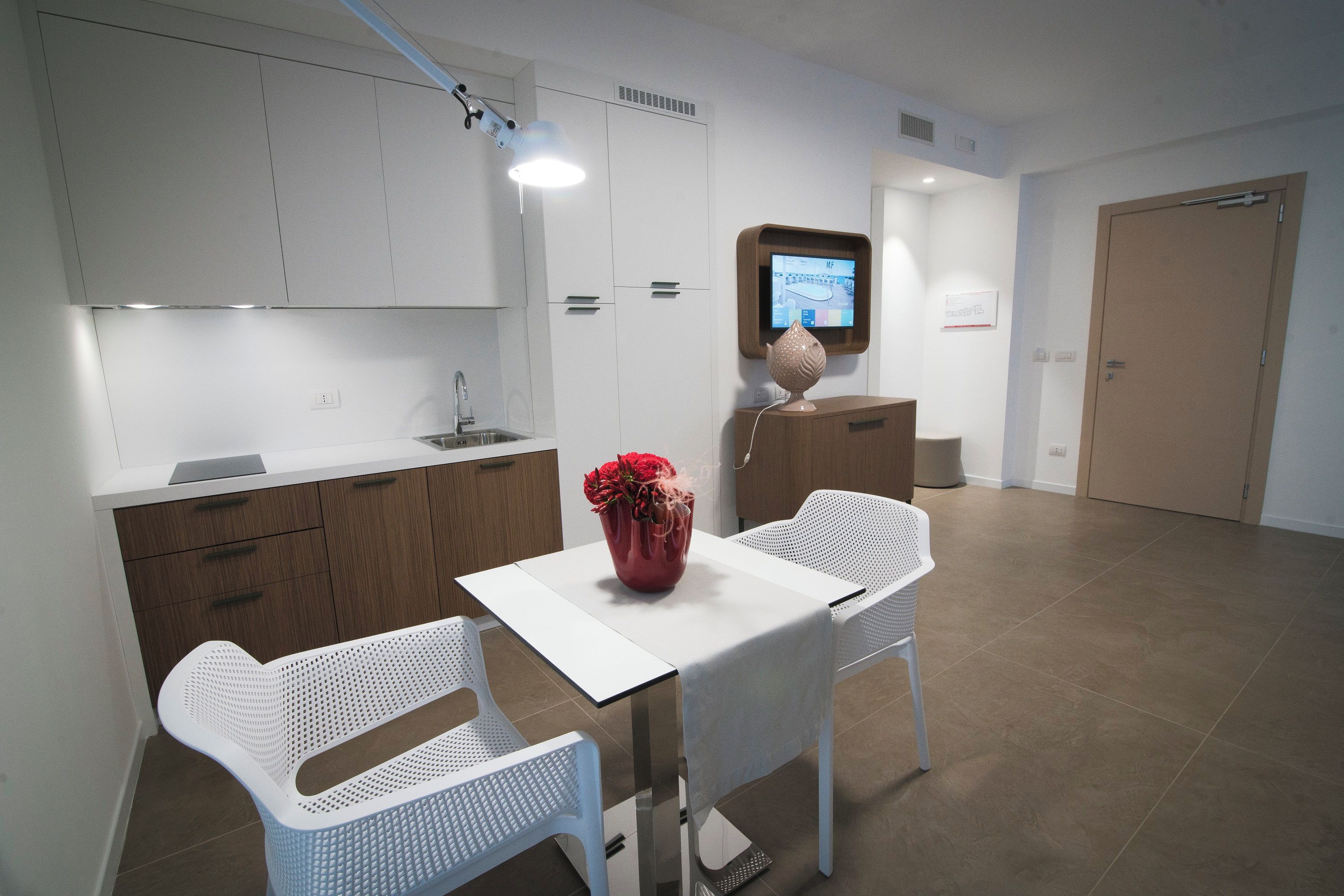 Une cuisine moderne avec des meubles blancs, un plan de travail en bois et une table blanche accompagnée de deux chaises. Un vase avec des fleurs rouges est posé sur la table.
