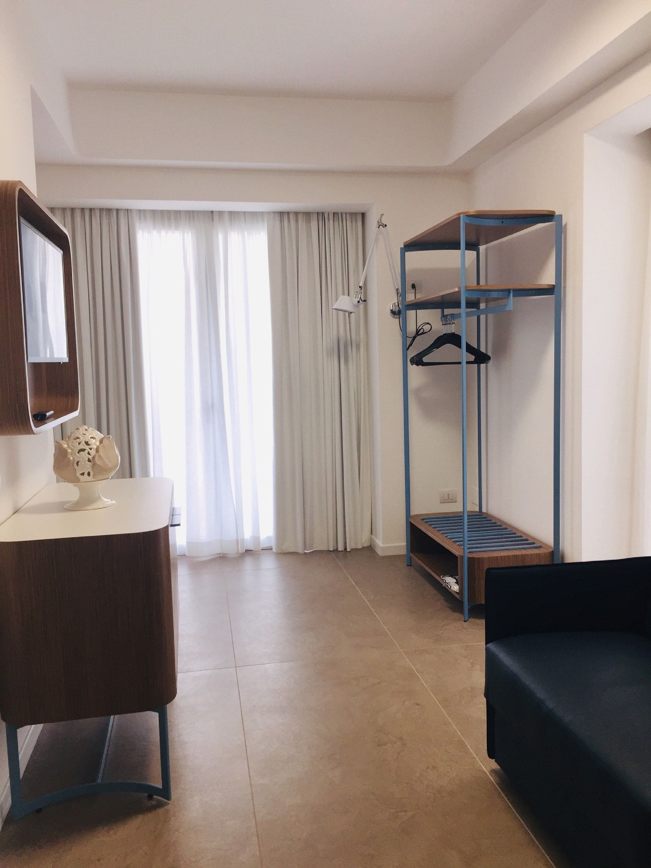 A hotel room with a wooden desk, a mirror, a bunk bed with a hanging rack, and a large window with curtains.