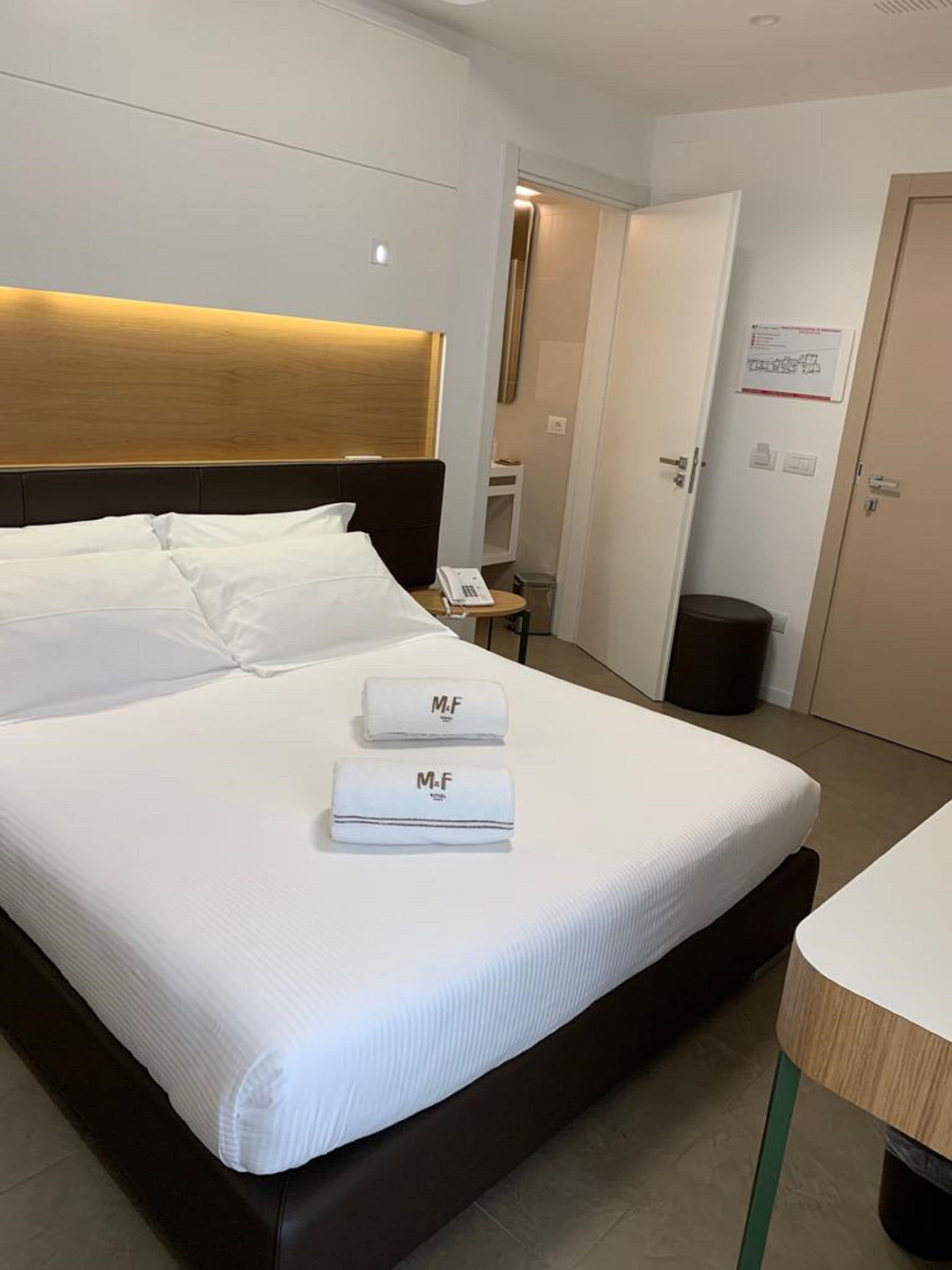 A hotel room with a large bed, two white towels with 'M.F' branding, a bedside table with a phone, and a door with a sign.