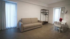 A modern living room with a beige sofa, a white dining table, and a white chair. There is a small shelf unit with a hanging rack and a floor lamp. - M&F Hotel (Gallipoli)
