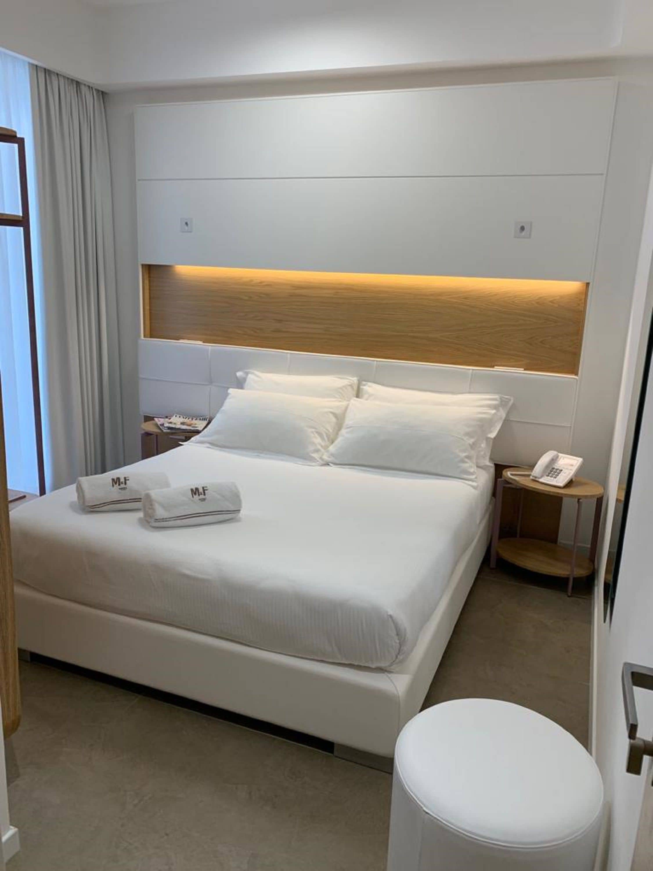 Un lit bien fait, avec du linge de lit blanc et deux serviettes ornées des initiales « M.F. », une table de chevet avec un téléphone et un petit pouf rond.