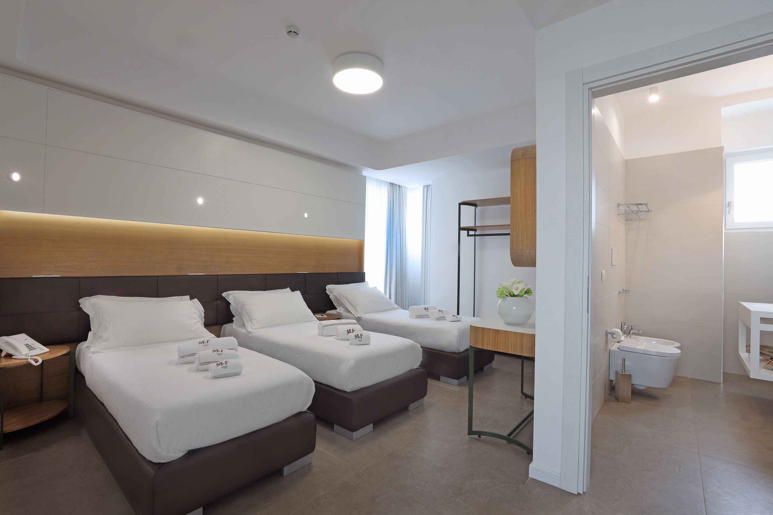 Habitación de hotel con dos camas, cabecero de madera, mesita de noche con teléfono, pequeño escritorio con lámpara y baño con ducha y lavabo.