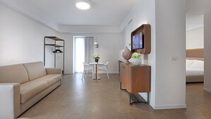 A modern hotel room with a beige sofa, a wooden cabinet with a TV, a dining area with a table and chairs, and a bed with white bedding. - M&F Hotel (Gallipoli)