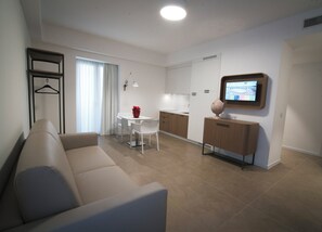 A modern living room with a sofa, a dining table with chairs, a kitchen area with cabinets, and a TV mounted on the wall. - M&F Hotel (Gallipoli)