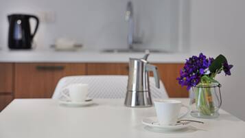 A white table with two white cups, a black coffee pot, and a clear glass vase with purple flowers.