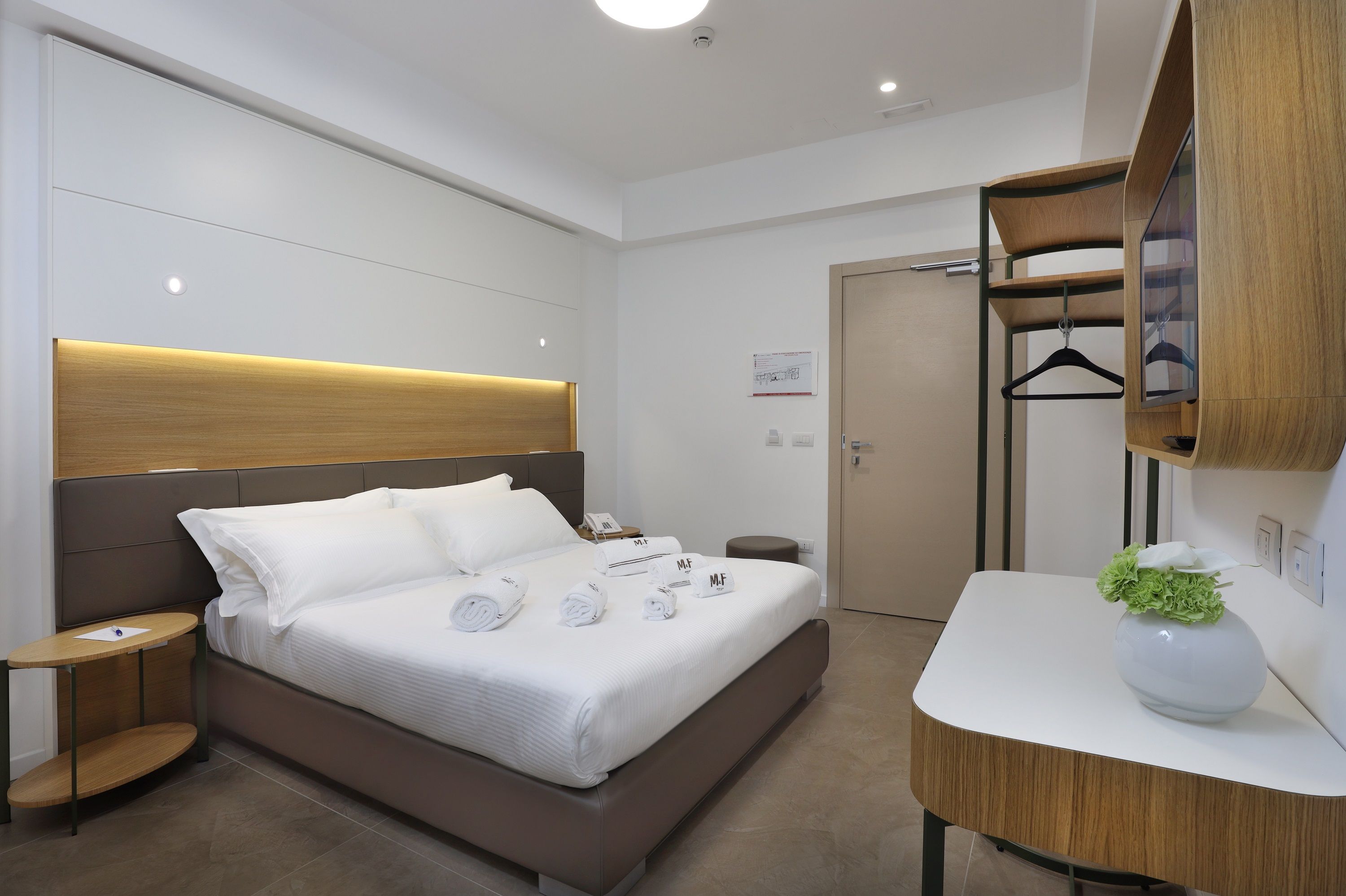 A modern hotel room with a large bed, bedside tables, a wooden headboard, a mirror, and a small plant on a bedside table.