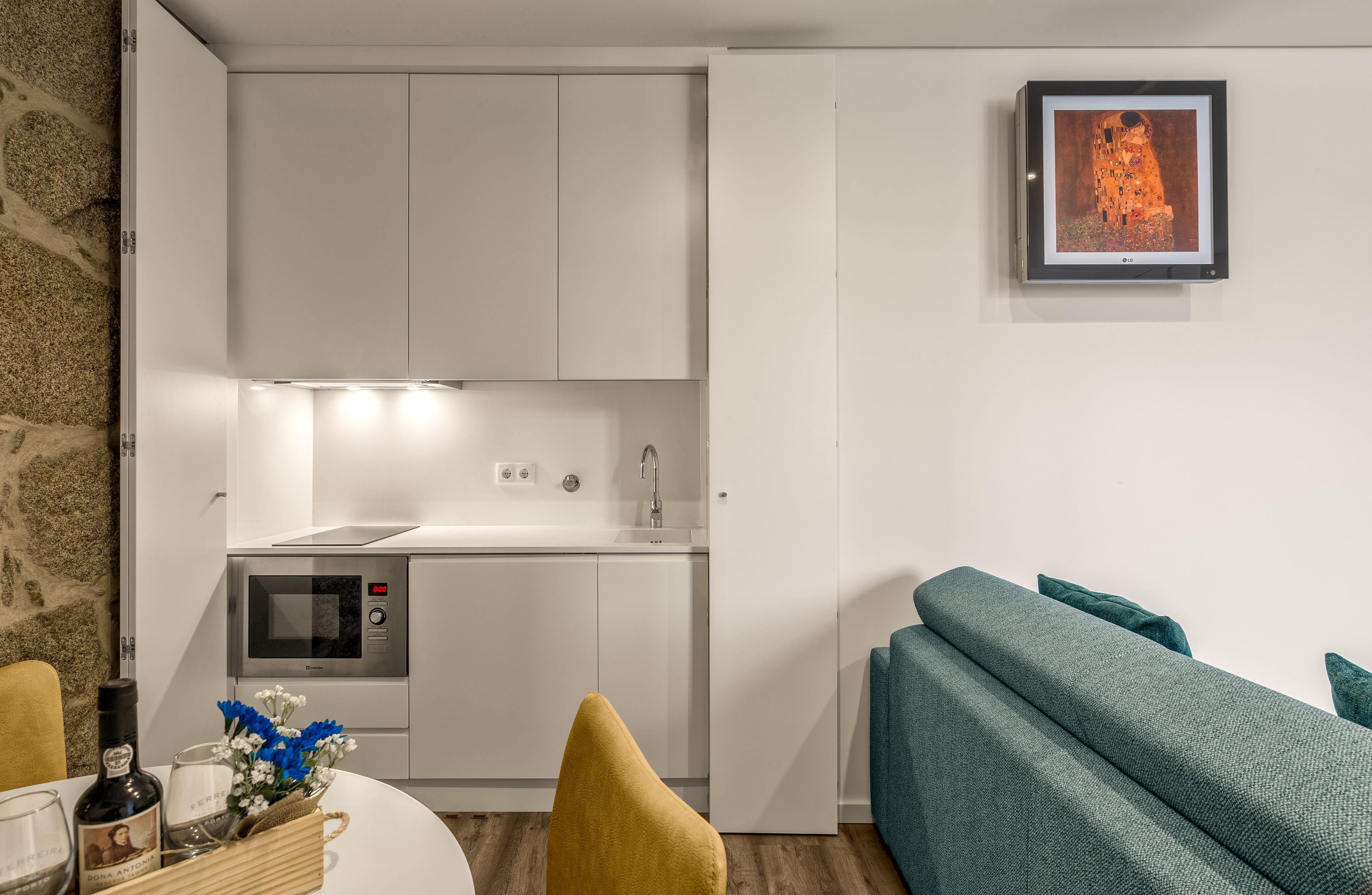 A modern kitchen with white cabinets, a built-in microwave, and a sink. There is a framed artwork on the wall and a green sofa in the living area.