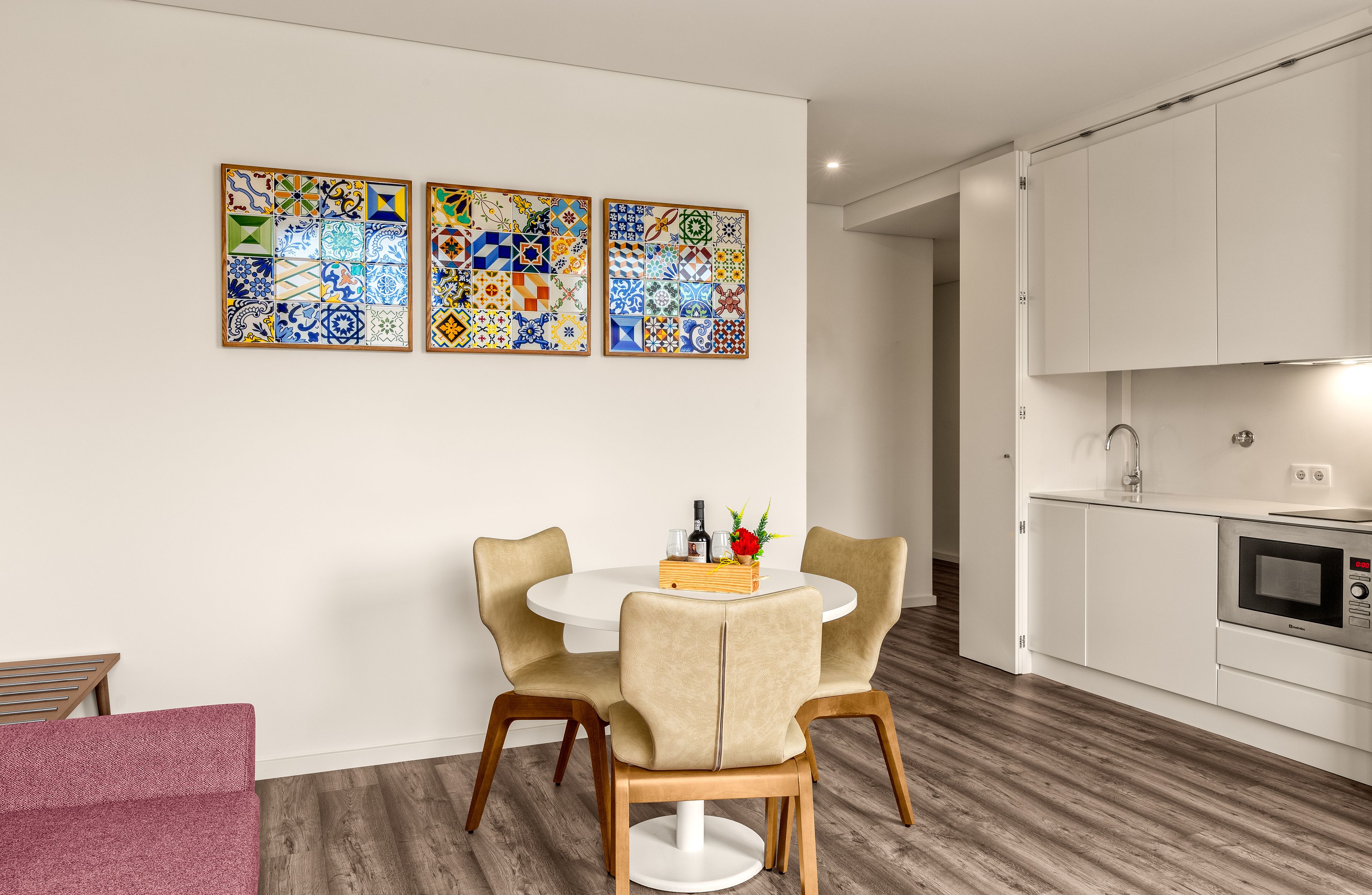 Eine moderne Küche mit weißen Schränken, einer Mikrowelle und einem Essbereich mit einem runden Tisch und zwei Stühlen. An der Wand befinden sich drei farbenfrohe Fliesenbilder.