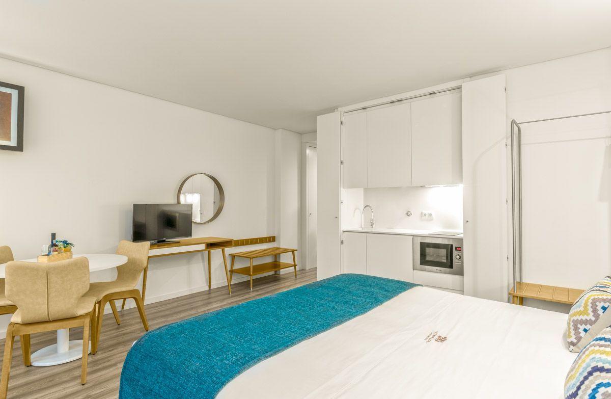 A hotel room with a bed, a desk with a chair, a small kitchen area, and a television.