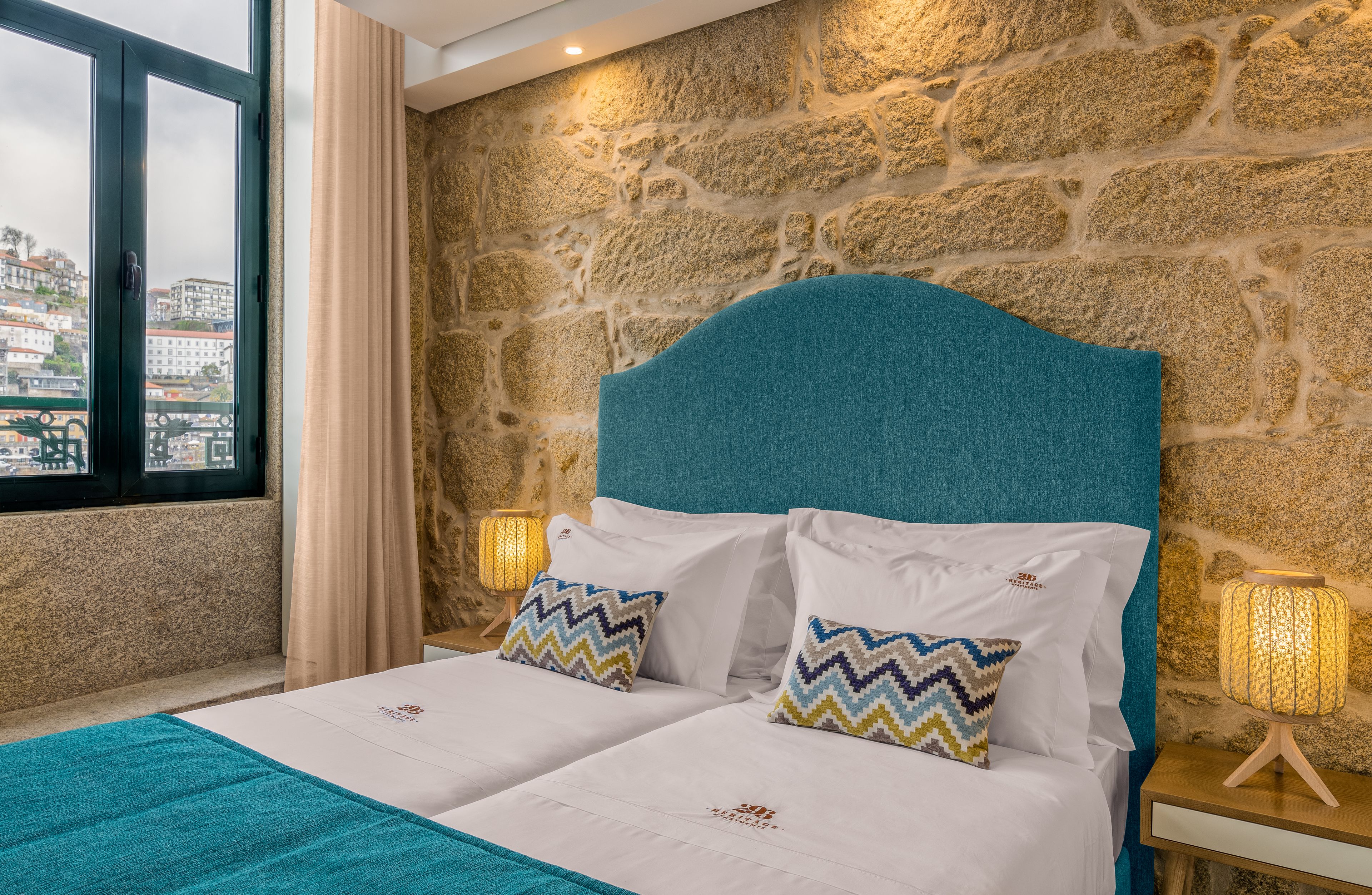 A hotel room with a stone wall, a bed with white bedding and patterned pillows, a view of a cityscape through a window, and a nightstand with a lamp.