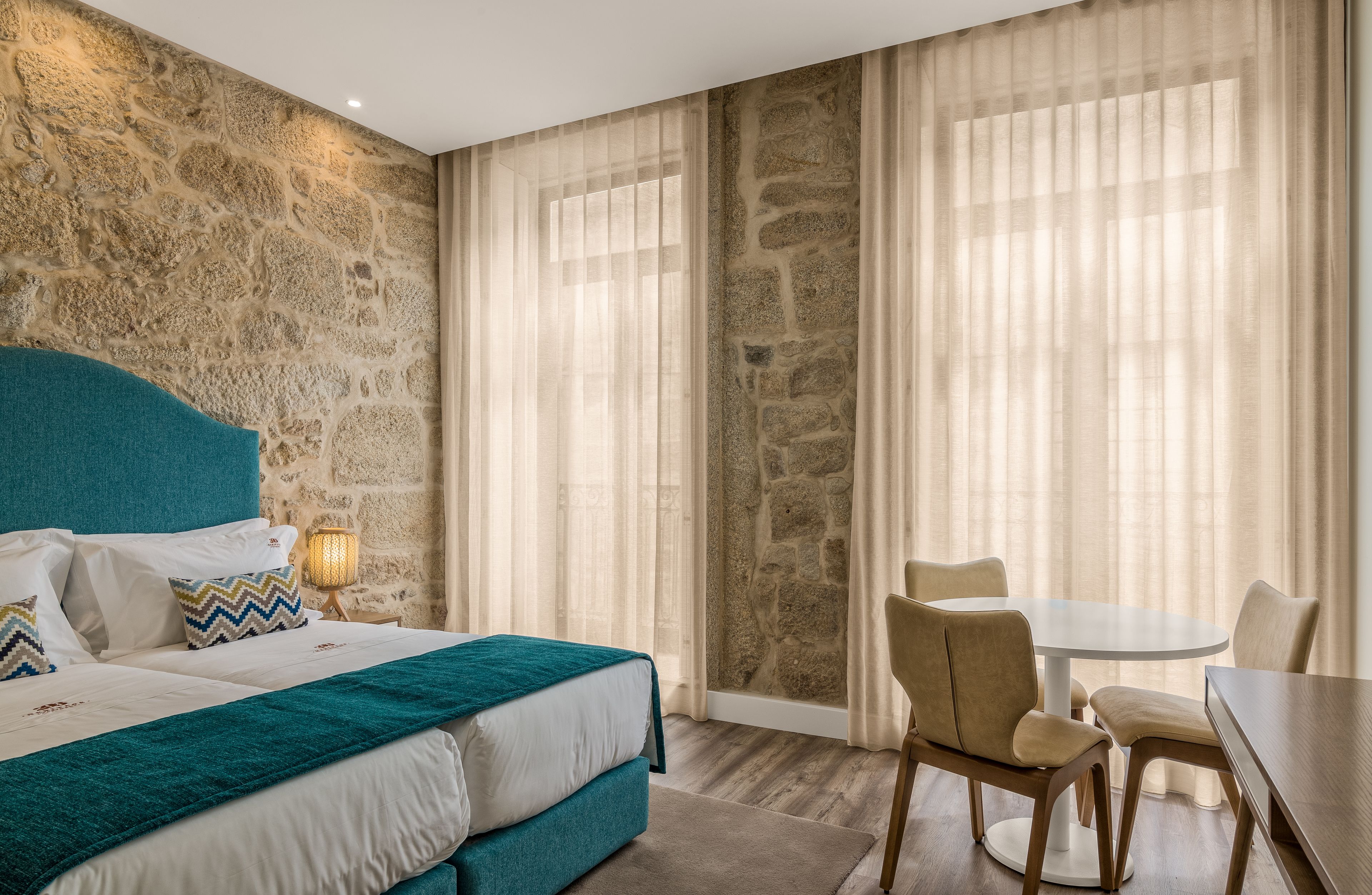 Une chambre d’hôtel avec un mur en pierre, un lit avec une literie de couleur turquoise, une table de salle à manger ronde et une chaise.