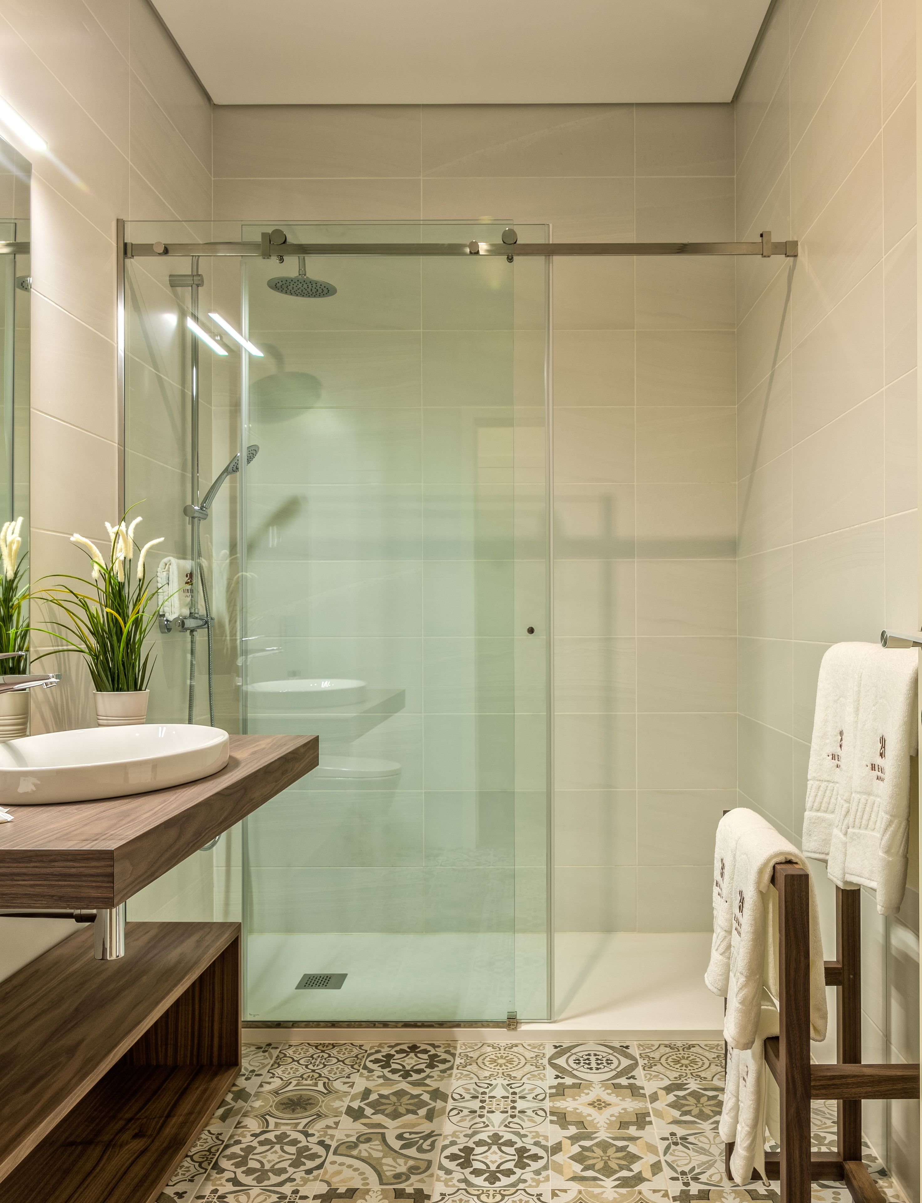 Un baño moderno con cabina de ducha de vidrio, tocador de madera con lavamanos y baldosas de suelo estampadas.