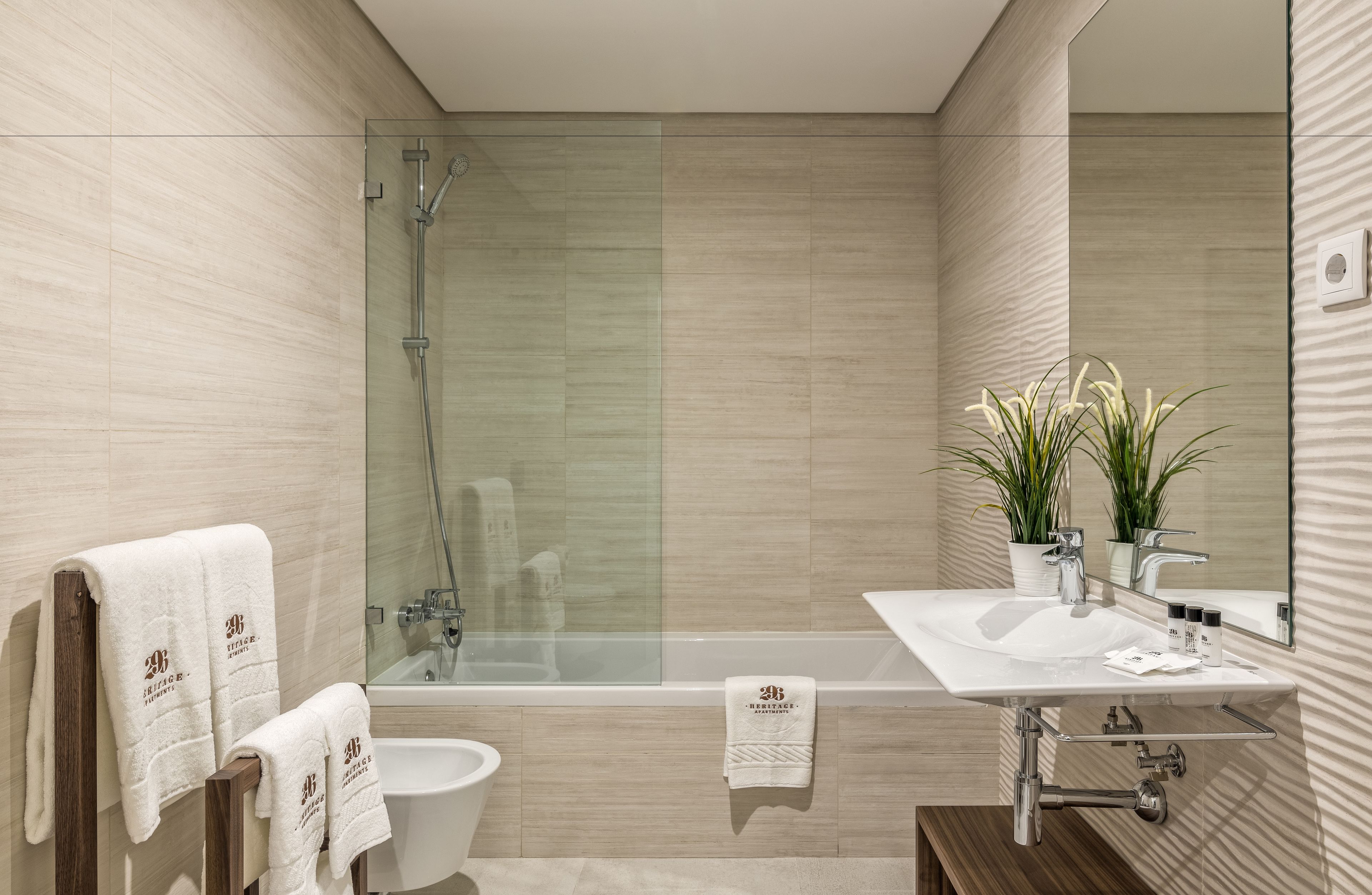 A modern bathroom with a glass-enclosed shower, a bathtub, a sink with a mirror, and a toilet. There are towels hanging on a wooden rack.
