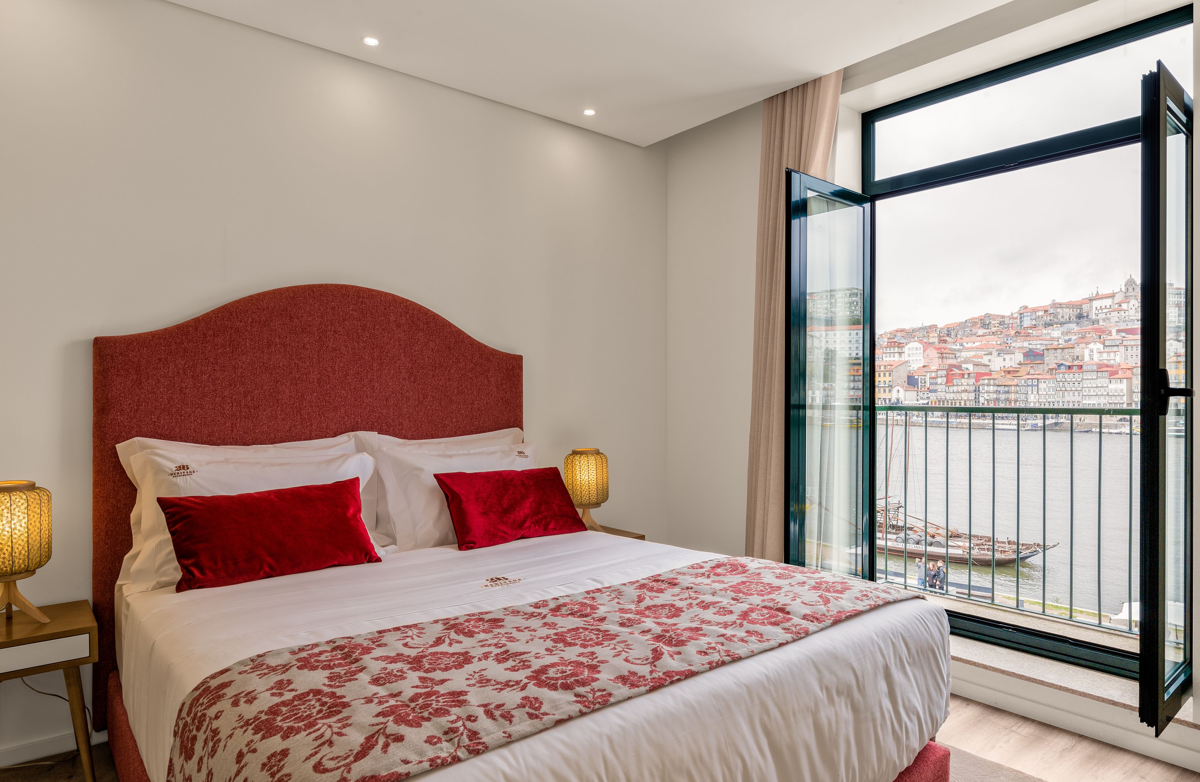 Quarto de hotel com uma cama grande, duas luminárias de cabeceira, uma janela com vista para a cidade e cabeceira vermelha.