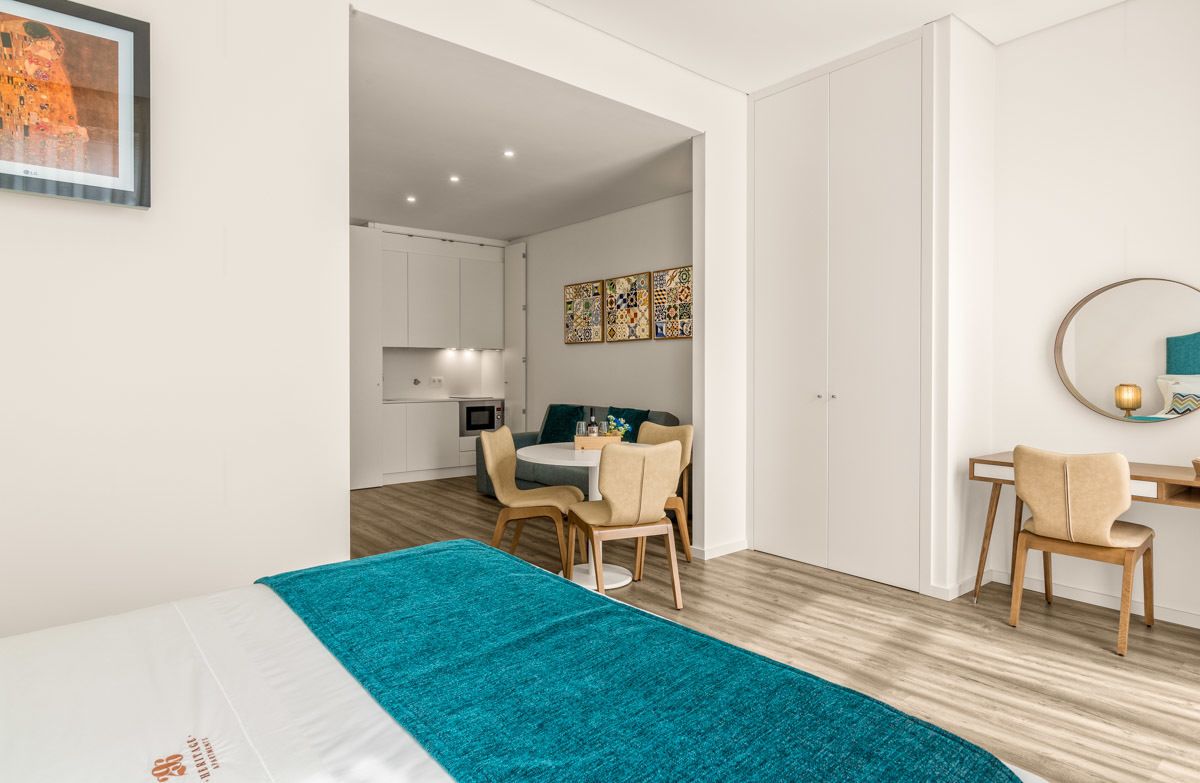 Ein modernes Hotelzimmer mit Bett, Schreibtisch, Stuhl, einem Esstisch mit Stühlen und einer durch eine offene Tür einsehbarer Küchenbereich.
