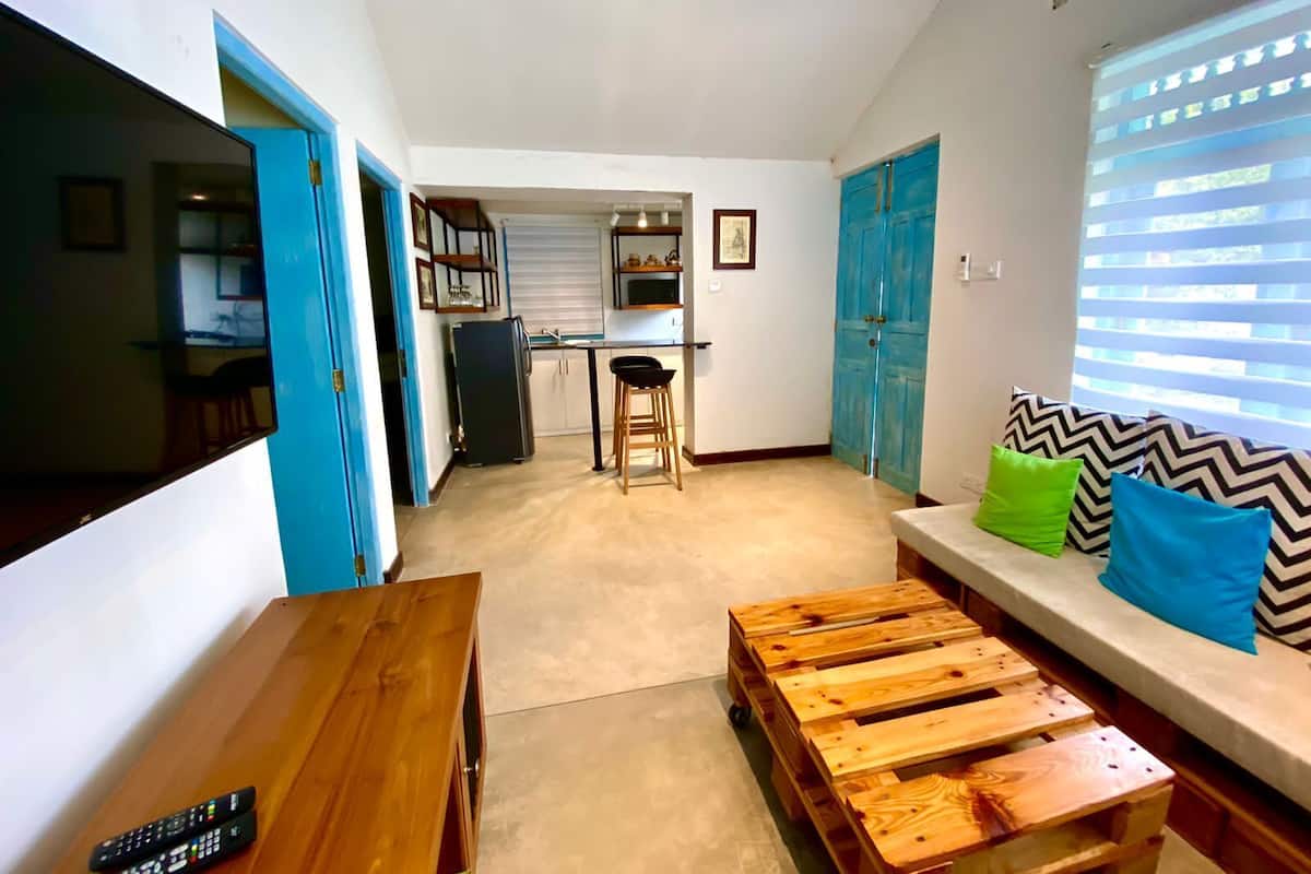 A living room with a wooden coffee table, a sofa with patterned cushions, a TV, and a kitchen area in the background.