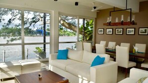 Ein modernes Wohnzimmer mit großem Fenster mit Blick auf ein Gewässer, ein weißes Sofa mit blauen Kissen, ein Holztisch und ein Essbereich mit Stühlen und einer Kristallleuchtre.