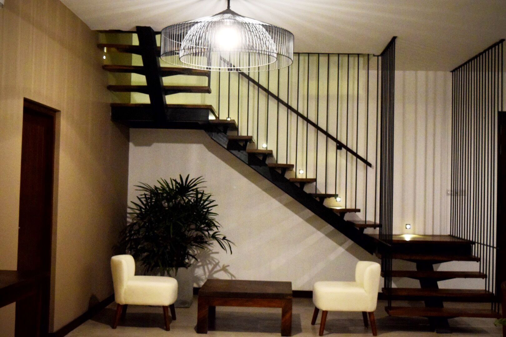 Una escalera moderna con barandilla negra, un candelabro y dos sillas blancas en una habitación con suelo de madera y una planta.