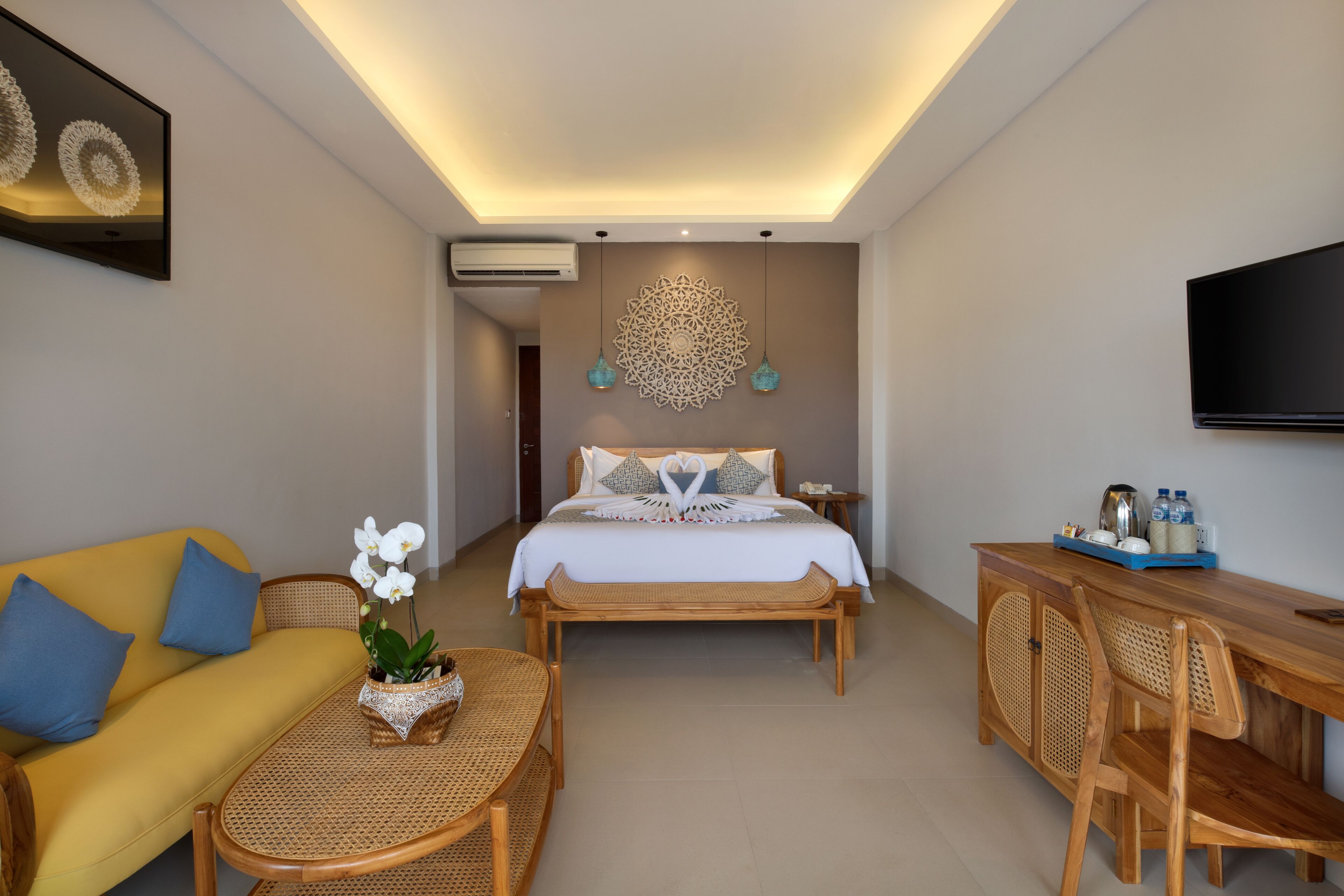 A hotel room with a bed, a sofa, a dining table, and chairs. There is a television on the wall and an air conditioning unit above the bed.